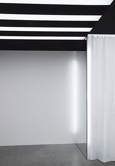 Deeper into the store, the horizontal and highly graphic lighting pattern is replaced with the glow of vertically-mounted fluorescent lights.  The draped, white gauze curtain of the fitting area is softly back-lit by the bare fluorescent tubes mounted at the front edges of the free-standing mirrored partitions.  Similar fluorescent tubes are placed on the opposite edge of each partition, along the perimeter wall.  The placement of the fixtures accentuates the freestanding character of the partitions, while their dimensions and finish match that of the partitions, contributing to the minimalist character of the design scheme.